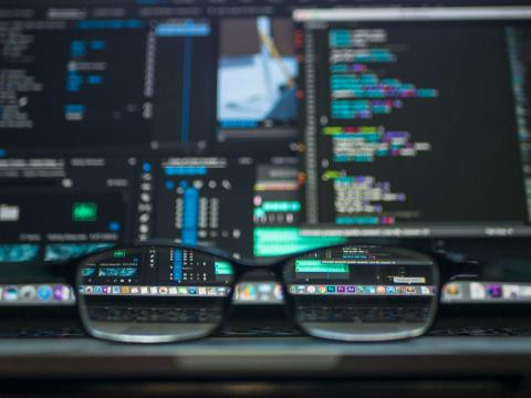 Glasses on a desk with screens in the background with code on them